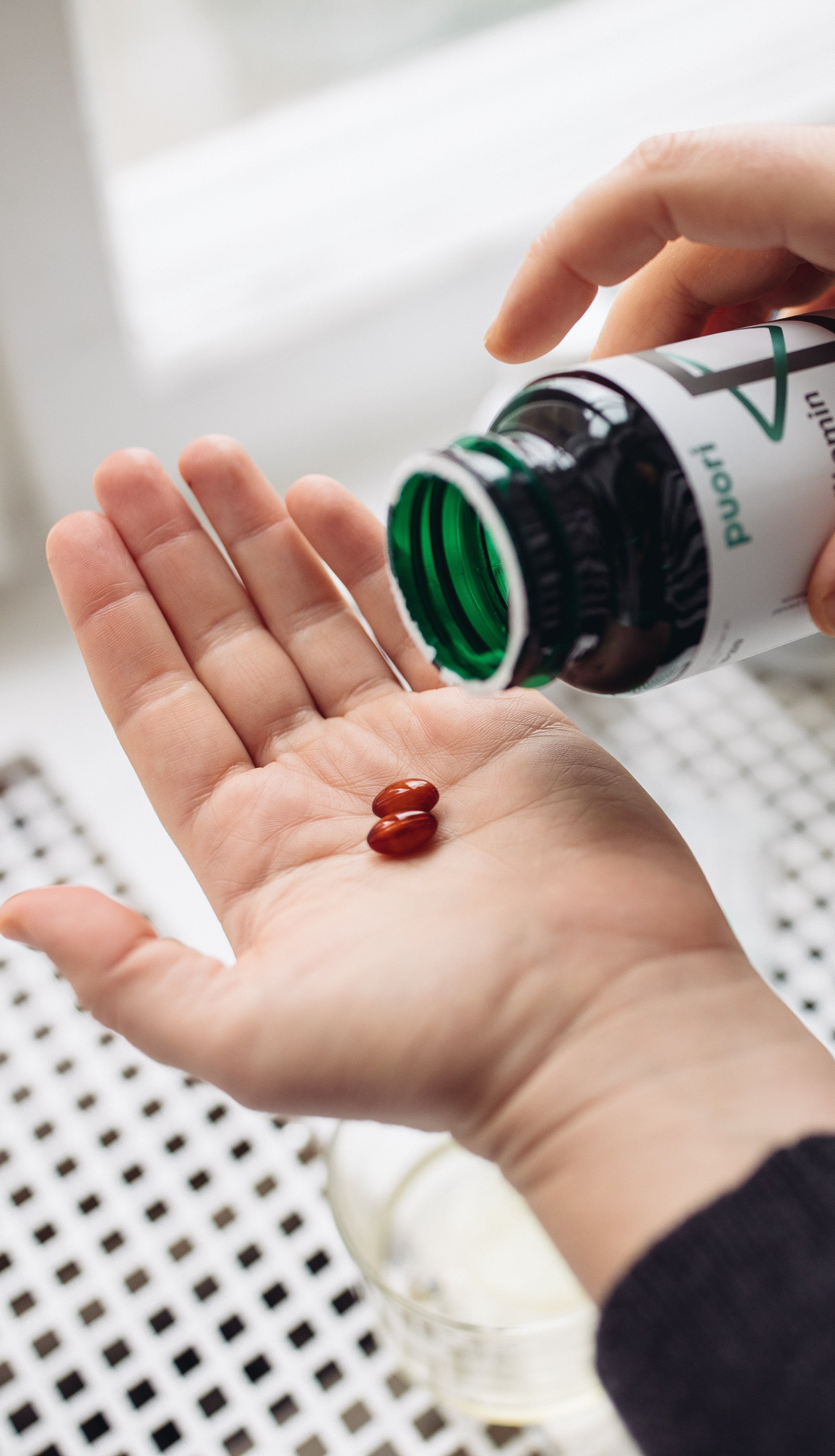 Close-up of two hands: one holding an open Puori vitamin bottle, tipping two brown softgel capsules into the open palm of the other hand.